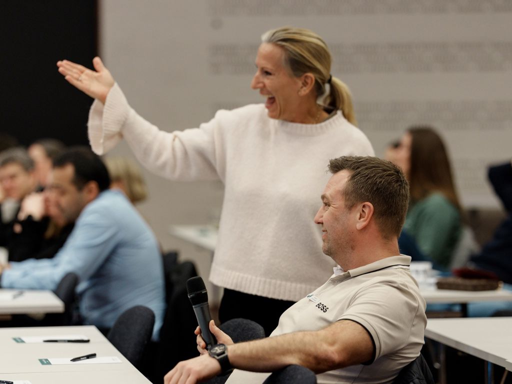Person står smilende ved siden af en person der sidder med en mikrofon