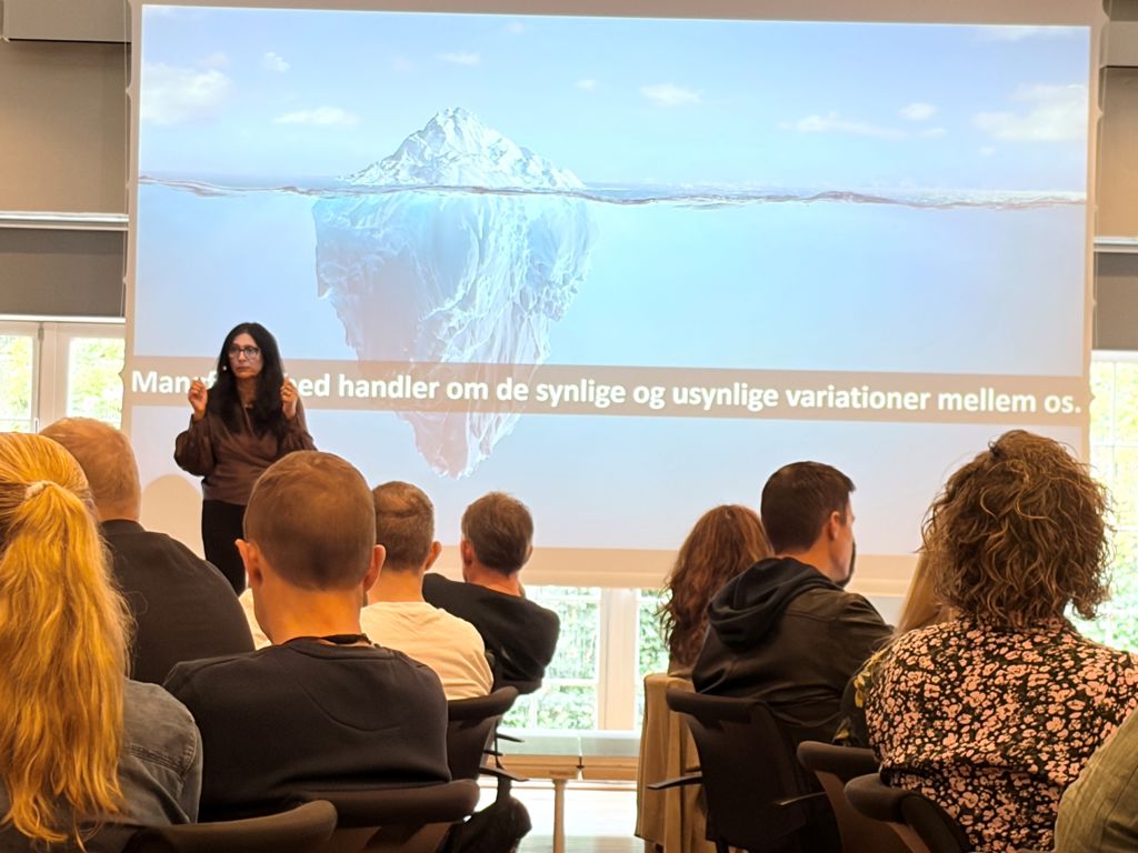 kvinde står foran præsentation og der sidder mennesker foran og lytter til hende.