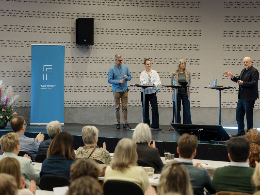 Fire personer på podiet i debat foran publikum med ryggen til.