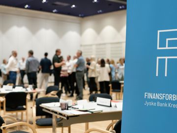 I forgrunden ses en roll-up med teksten Finansforbundet Jyske Bank Kreds. I baggrunden ses en større forsamling tillidsrepræsentanter i dialog. 