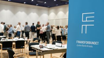 I forgrunden ses en roll-up med teksten Finansforbundet Jyske Bank Kreds. I baggrunden ses en større forsamling tillidsrepræsentanter i dialog. 