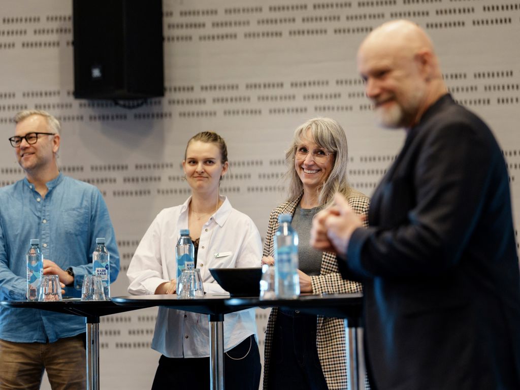 4 smilenden personer stående foran høje borde
