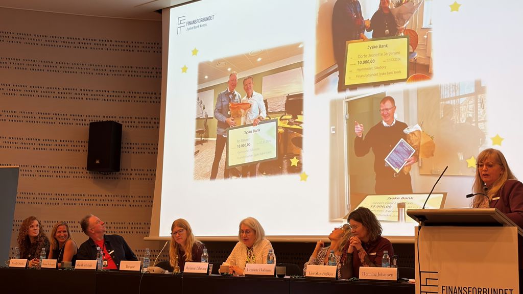 Person på talerstolen. I baggrunden præsentation på storskærm samt 6 personer siddende i panel.
