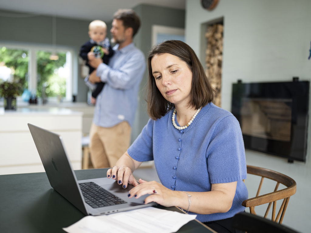 En mor sidder ved sin computer mens far og baby er i baggrunden